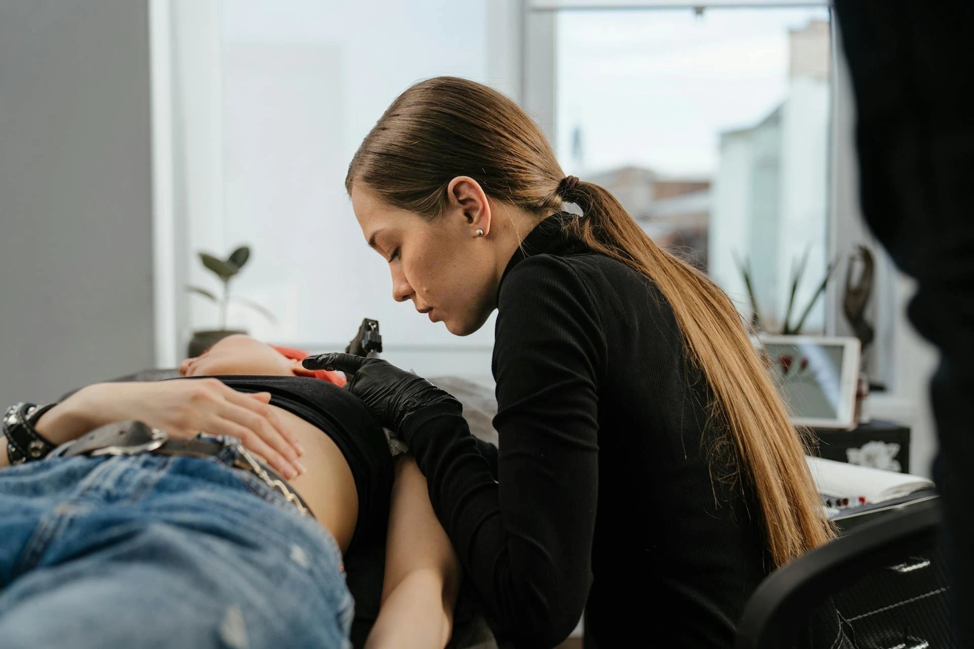 Tatoueuse professionnelle assise, vêtue d'une tenue noire et gants latex noir, en train de réaliser un tatouage sur un client allongé, dans un studio de tatouage équipé et éclairé.
